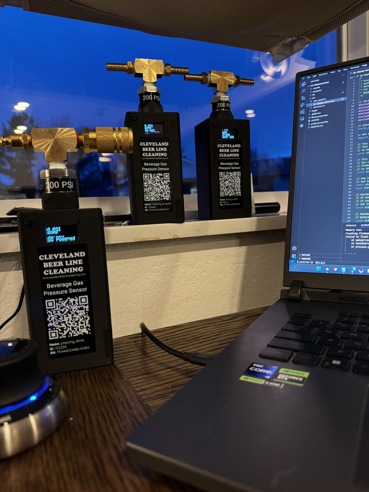 Three early Barkeep beverage gas pressure sensors on a windowsill showing live PSI readings, next to a laptop running the sensor firmware in a code editor.